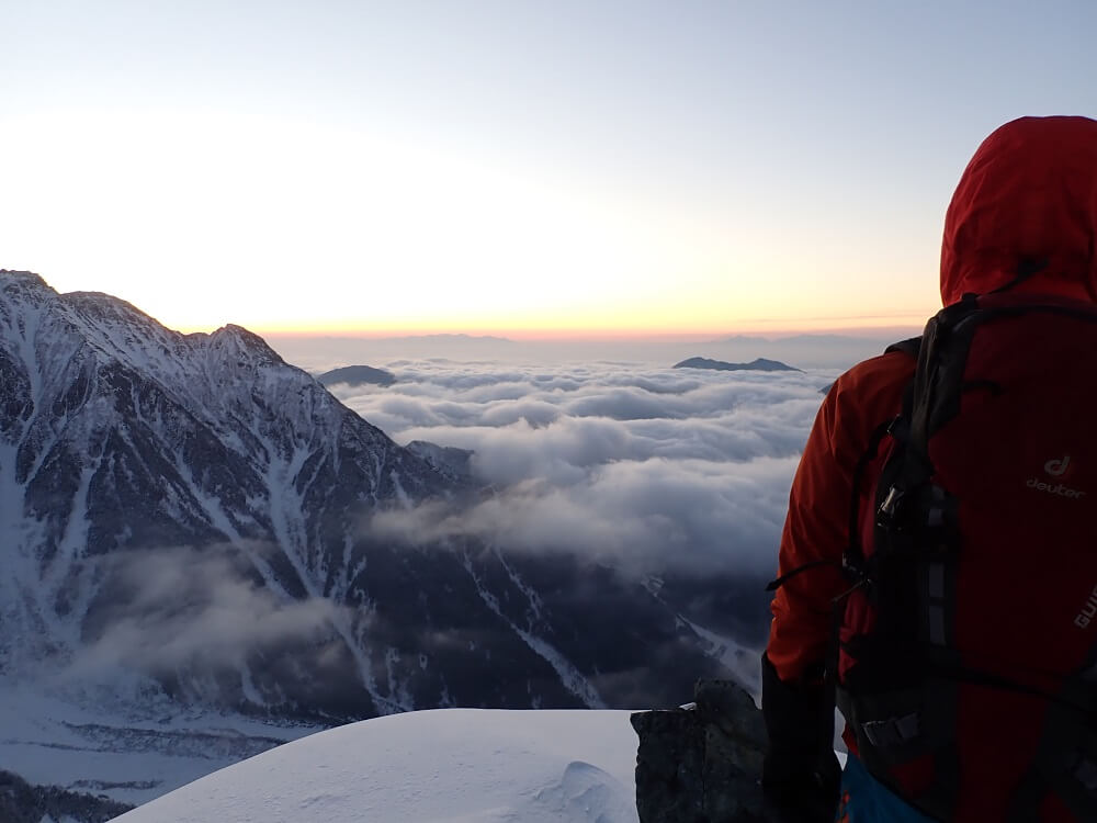 Hiker in red parka standing on a snowy mountain top looking towards the horizon, photographed with the Olympus TG-6